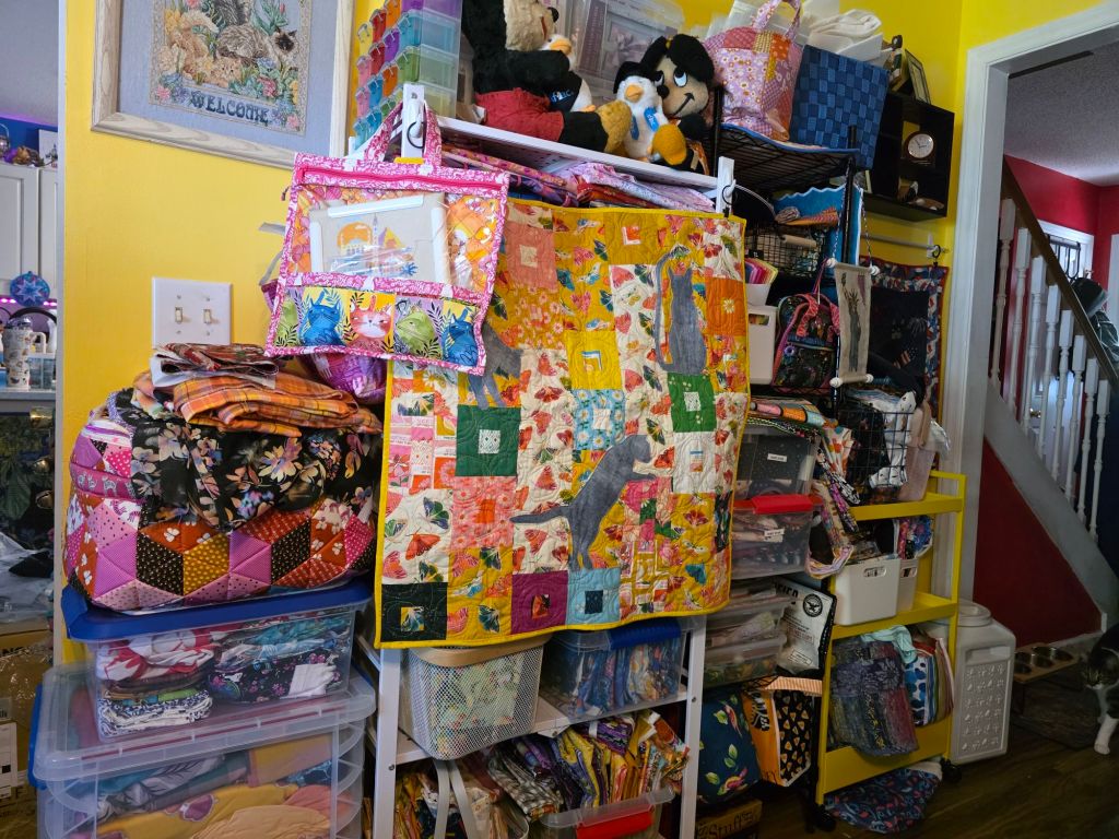 A colorful craft room filled with shelves of fabric, quilting supplies, and various bags. There is a vibrant quilt hanging on the shelf, along with decorative items and plush toys. The walls are painted yellow, and a staircase can be seen in the background.