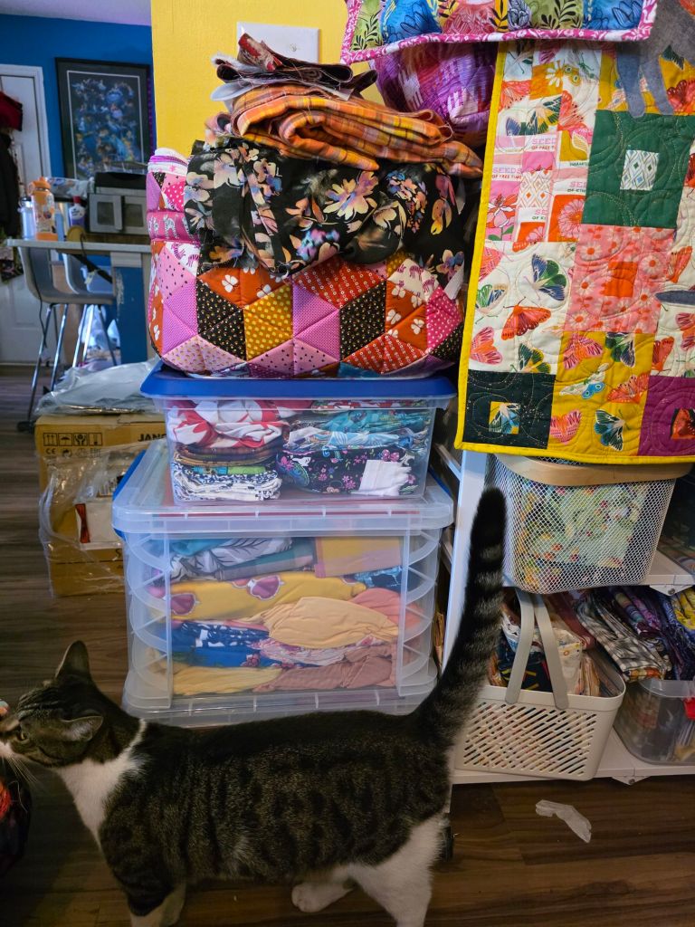 A cluttered workspace featuring a stack of colorful quilts and fabrics atop storage containers, with a tabby cat walking in front.