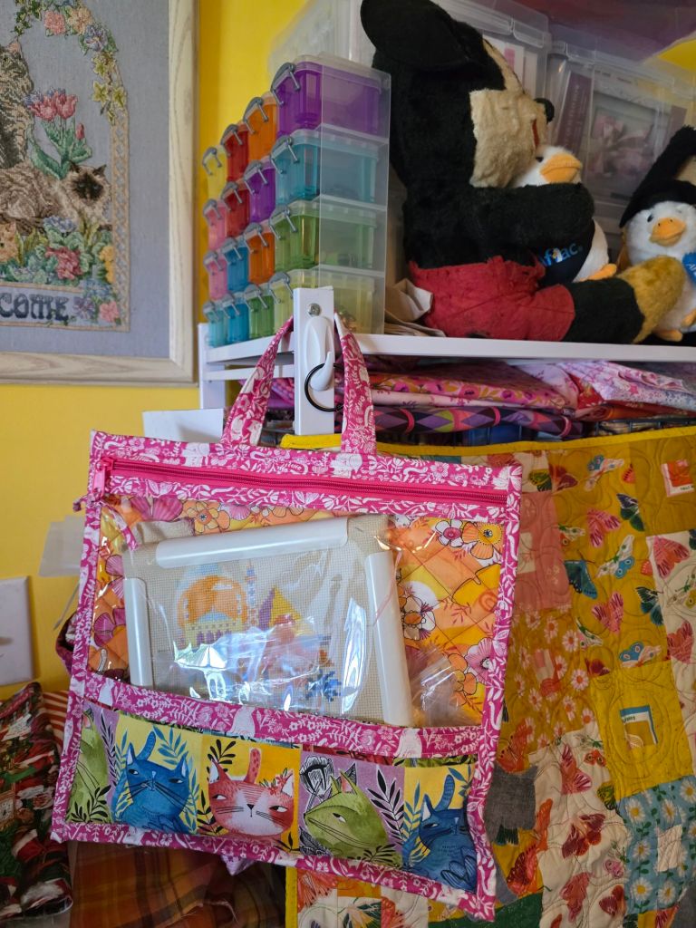 A colorful wall organizer with a transparent pocket displaying crafting supplies, including fabric, threads, and a frame, hanging next to stuffed animals and storage boxes in a bright room.