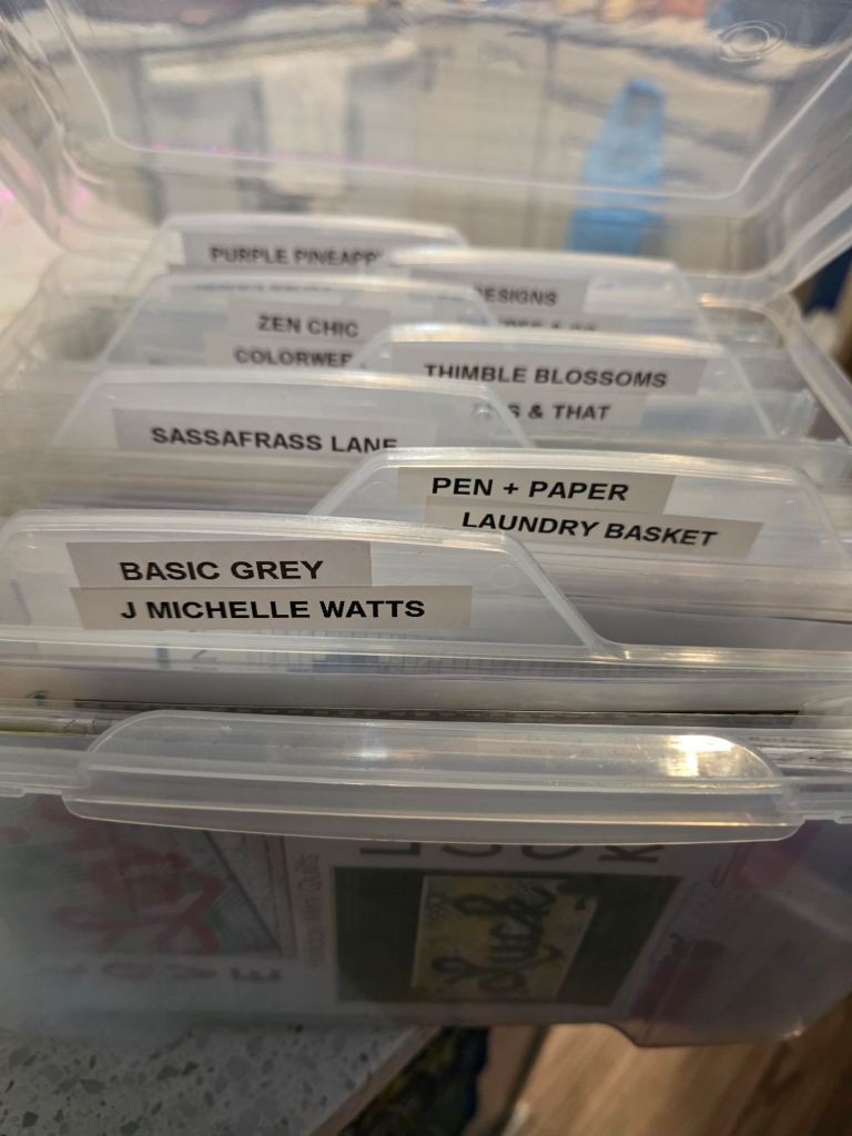 A clear plastic container filled with labeled dividers for various crafting supplies, including 'Purple Pineapple', 'Zen Chic', and 'Basic Grey'.