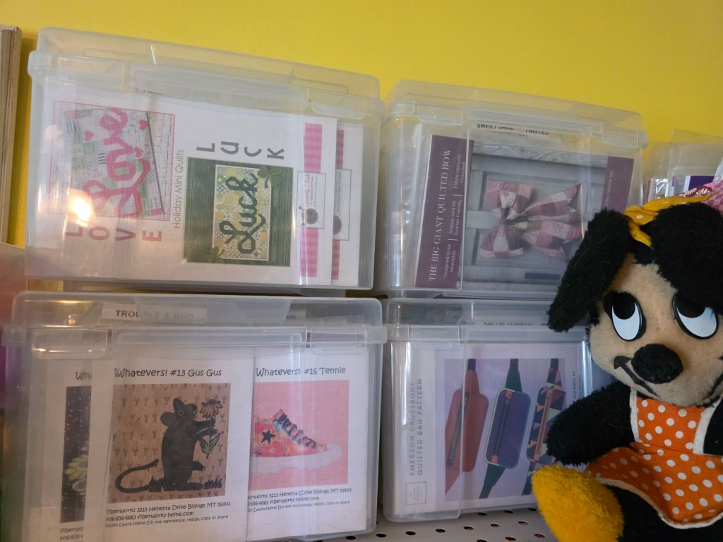Storage boxes containing quilting patterns and projects with labels like 'Love,' 'Luck,' and 'The Big Giant Quilted Box.' A plush dog toy wearing an orange dress is positioned next to the boxes.