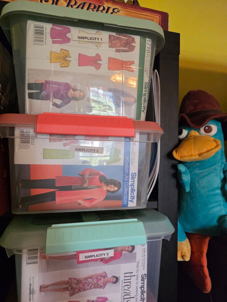 Three storage bins labeled 'Simplicity 1', 'Simplicity 2', and 'Simplicity 3', containing sewing patterns for clothing, with a plush character resembling a blue platypus in the background.