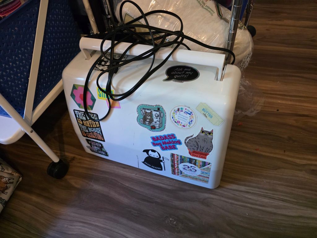 A white sewing machine covered in colorful stickers, with a power cord coiled on top, sitting on a wooden floor near fabric and other craft materials.