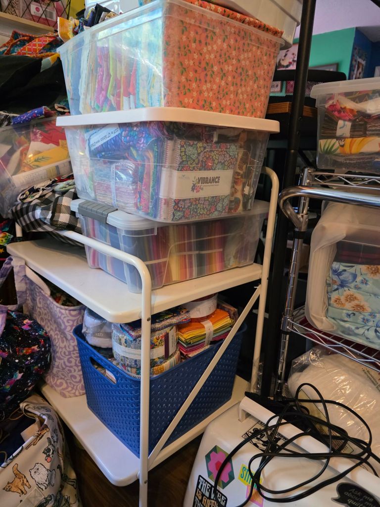 A cluttered workspace featuring stacked transparent storage bins filled with colorful fabrics and sewing supplies, organized on a white shelf.