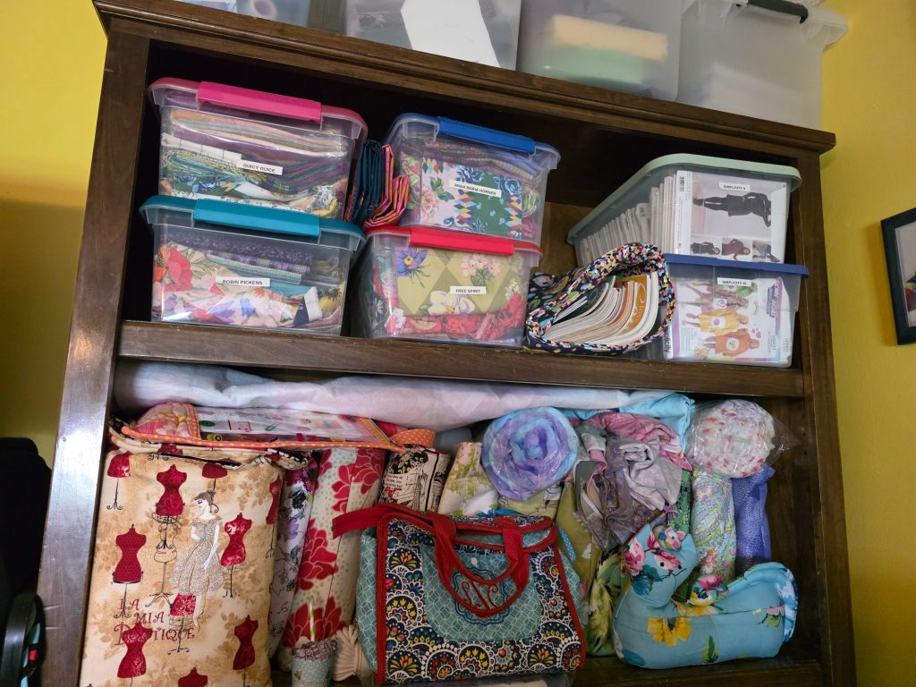 A wooden shelf filled with colorful storage containers, fabric rolls, and sewing accessories, set against a yellow wall.