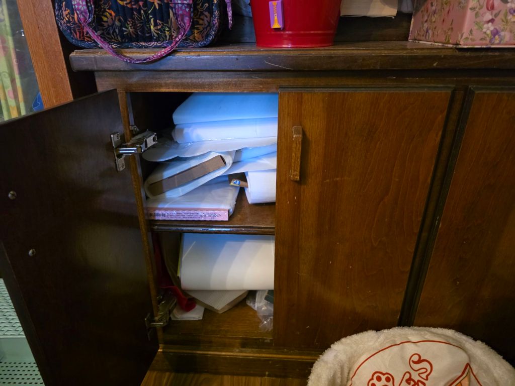 Open wooden cabinet with a clutter of assorted materials, including stacked paper and boxes, partially filled with organizational items.
