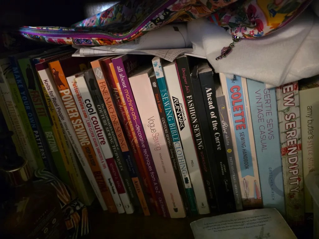 A collection of sewing books stacked on a shelf, with a colorful zipper pouch at the top and some loose papers tucked among the books.