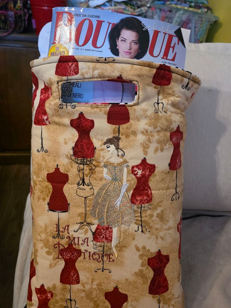 A decorative fabric bag featuring a design with vintage dress forms and the text 'La Mia Boutique,' holding a sewing magazine titled 'BOUTIQUE' with a woman's portrait on the cover.