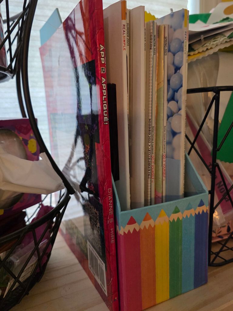 A colorful pencil-shaped container holding various magazines and a book titled 'APP 2 APPLIQUE!' and 'DIANNE'S HIRE' on the spine.