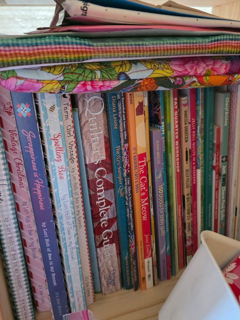 A close-up view of a bookshelf displaying a variety of colorful quilting and craft books, alongside folders, fabrics, and containers.
