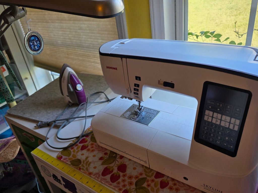 A Janome Skyline sewing machine on a table, with an iron in the background and a window with shades providing natural light.