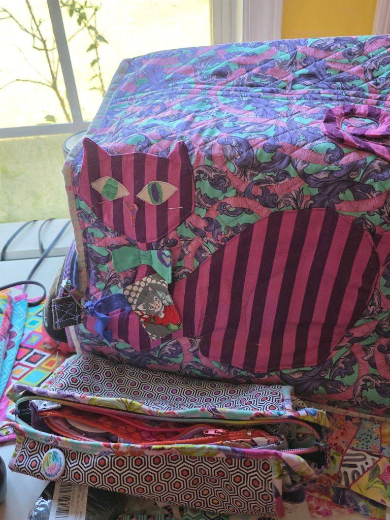 A colorful sewing machine cover designed with a whimsical cat motif, featuring vibrant patterns and colors, placed on a table beside various fabric bags.