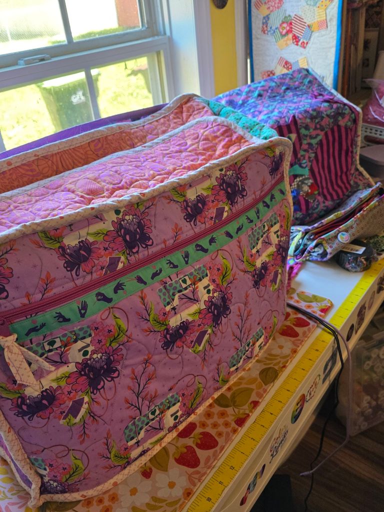 Two colorful sewing machine bags with floral patterns, placed on a table near a window. A ruler and additional sewing supplies are visible on the table.