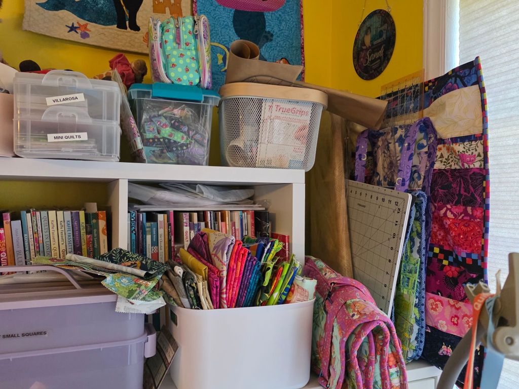 A cluttered craft workspace featuring shelves filled with colorful fabric, books on quilting, and storage bins labeled for mini quilts and small squares.