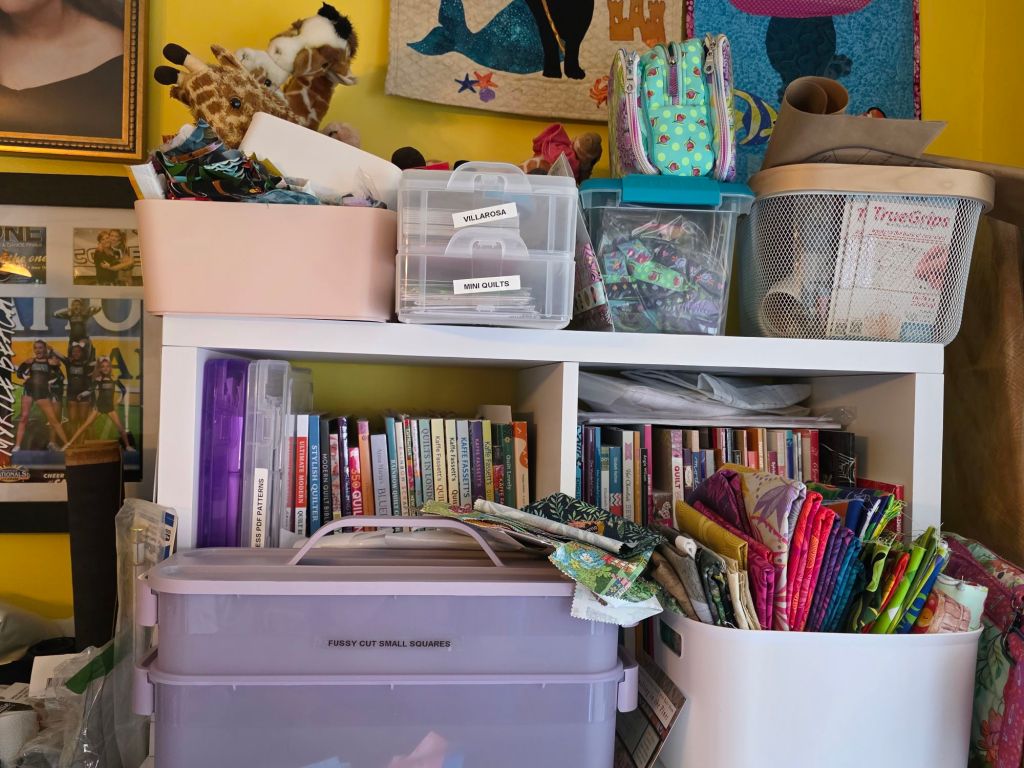 A cluttered shelf filled with various containers of fabric, quilting books, and craft supplies, set against a bright yellow wall.