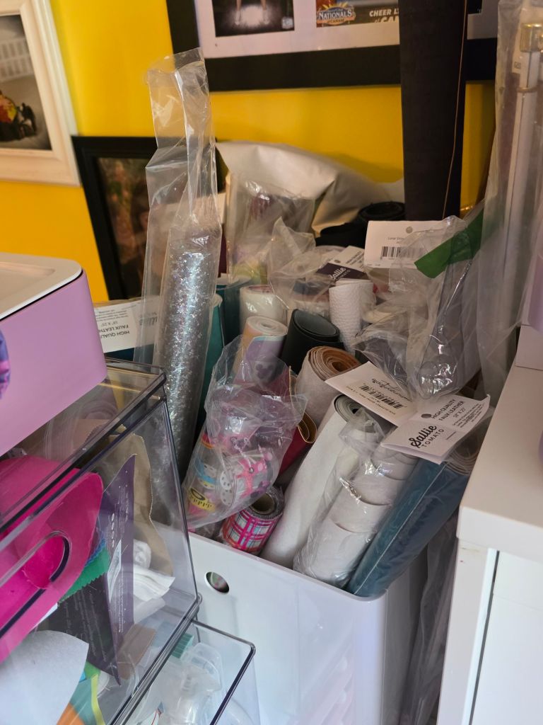 A cluttered storage area filled with various rolls of paper, stickers, and craft supplies, surrounded by plastic packaging and colored materials.