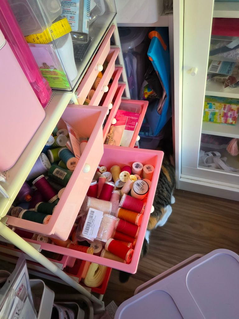 A craft organization unit with pink drawers filled with various spools of thread in different colors, along with notions and supplies. A glimpse of a cat is seen in the background.