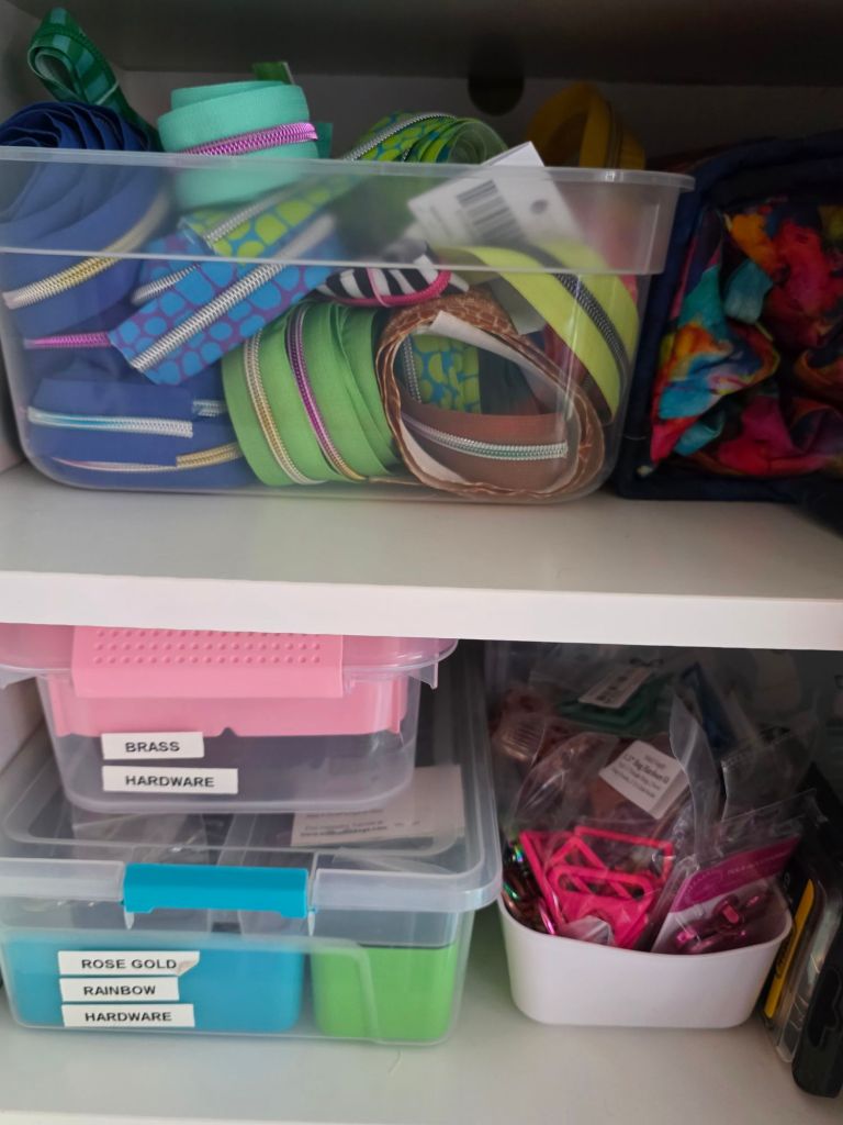 Organized storage shelves with bins containing various zippers, hardware, and craft supplies in different colors.