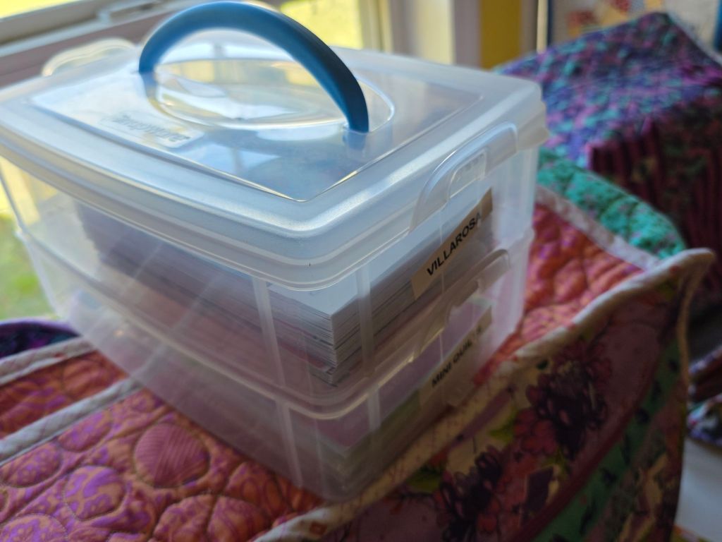 Clear storage container with a blue handle, filled with labeled cards, placed on a colorful quilt.