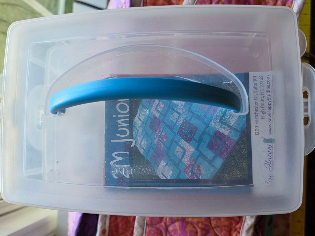 A clear plastic storage container with a blue handle, containing a booklet titled 'M Junior' and colorful patchwork fabric patterns visible inside.