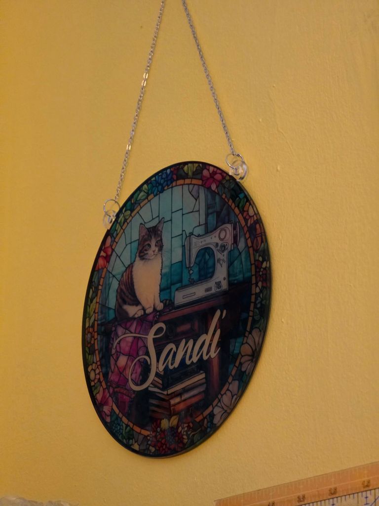 Colorful stained glass sign featuring a cat and a sewing machine with the name 'Sandi' written in the center, hanging on a yellow wall.