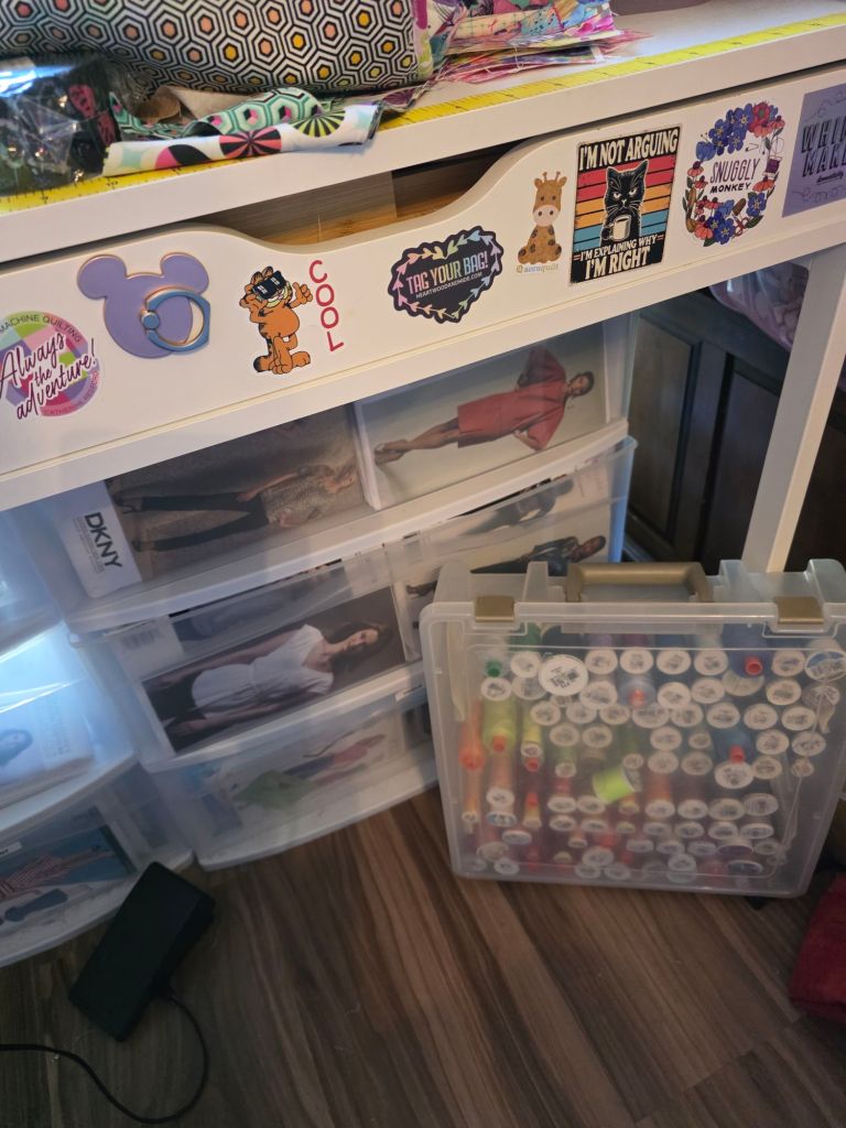 A cluttered workspace featuring a drawer with colorful stickers, including cartoon characters and phrases, alongside transparent storage bins filled with images and craft supplies.