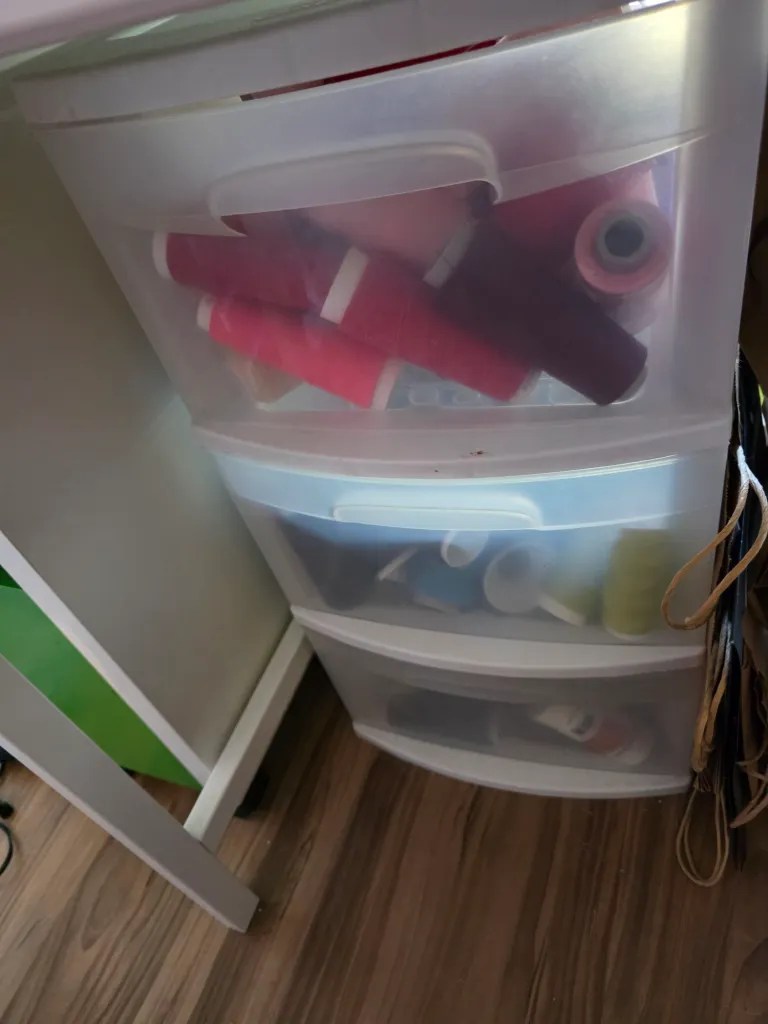 A stack of clear plastic storage drawers filled with various spools of thread in shades of red, pink, and blue, situated next to a wooden floor.