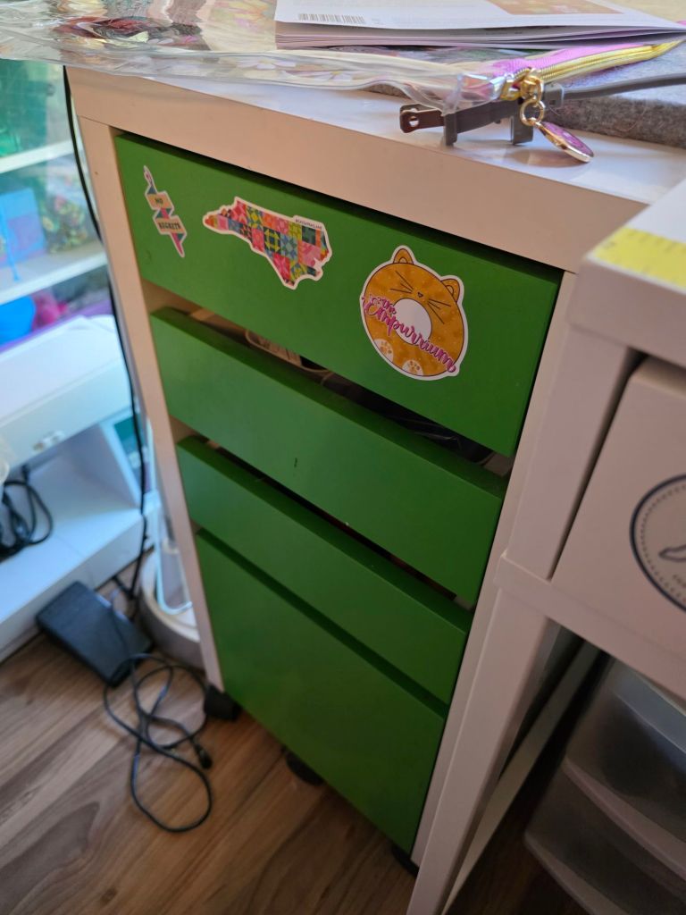 A green three-drawer storage unit with colorful stickers on the front, including a state outline and a cartoon cat. A transparent pouch and other items are on top of the unit.