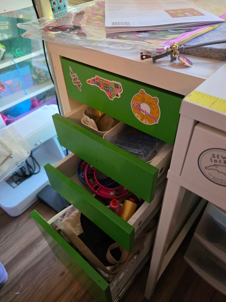 A close-up view of a green dresser with several open drawers, displaying various craft supplies including threads, tools, and colorful items, with decorative stickers on the front of the drawers.