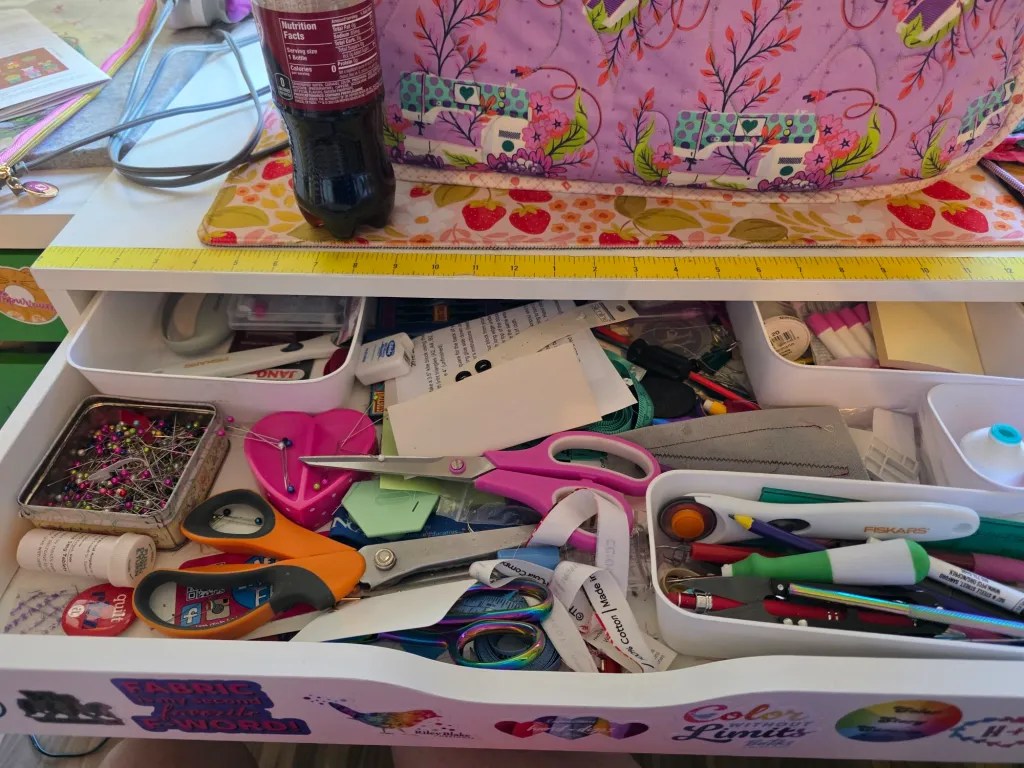 A cluttered craft drawer filled with various supplies including scissors, pens, pins, papers, and a bottle of soda on top.