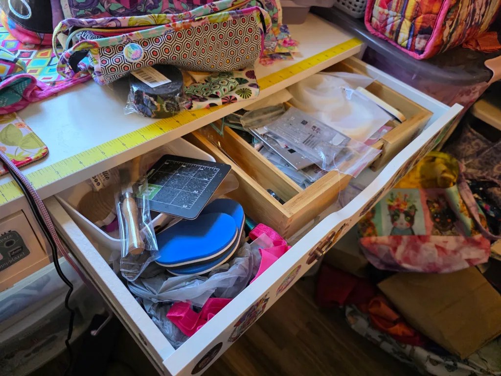 A cluttered craft drawer filled with various tools and materials, including measuring tools, fabric pieces, and accessories, set against a colorful, patterned workspace.