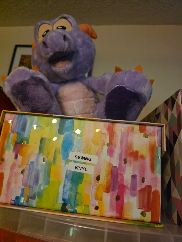 A plush purple dragon toy sitting on top of a colorful box labeled 'SEWING VINYL', featuring a vibrant abstract watercolor design.