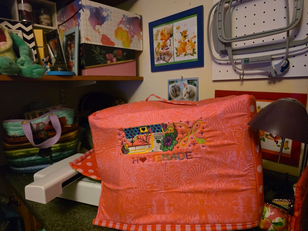 A colorful storage bag with an embroidered design that says 'Homemade', placed on a craft table surrounded by various craft supplies, boxes, and artwork on the walls.