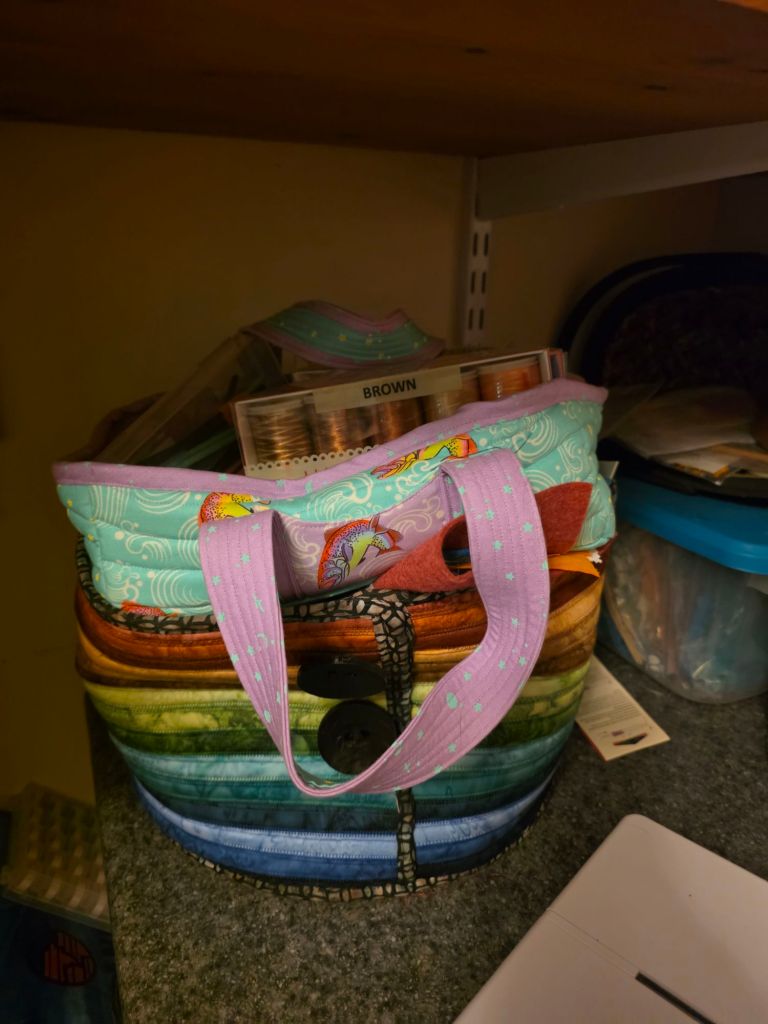 A colorful fabric bag with rainbow stripes and a pink handle, filled with craft supplies and spools of thread, sitting on a countertop.