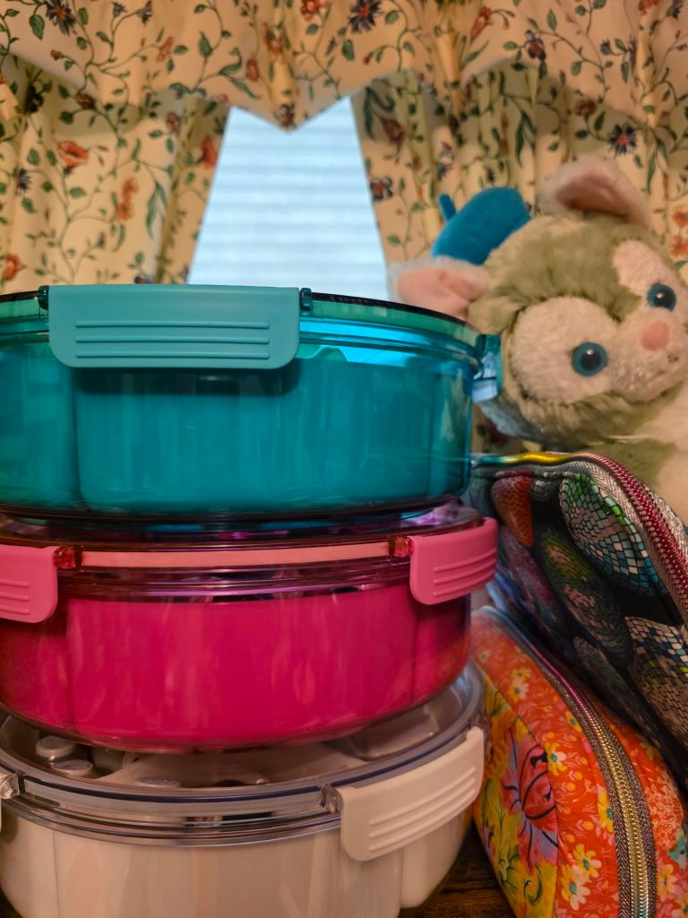 Stacked food storage containers in teal, pink, and white, beside a colorful zippered pouch and a plush toy, with a floral curtain in the background.
