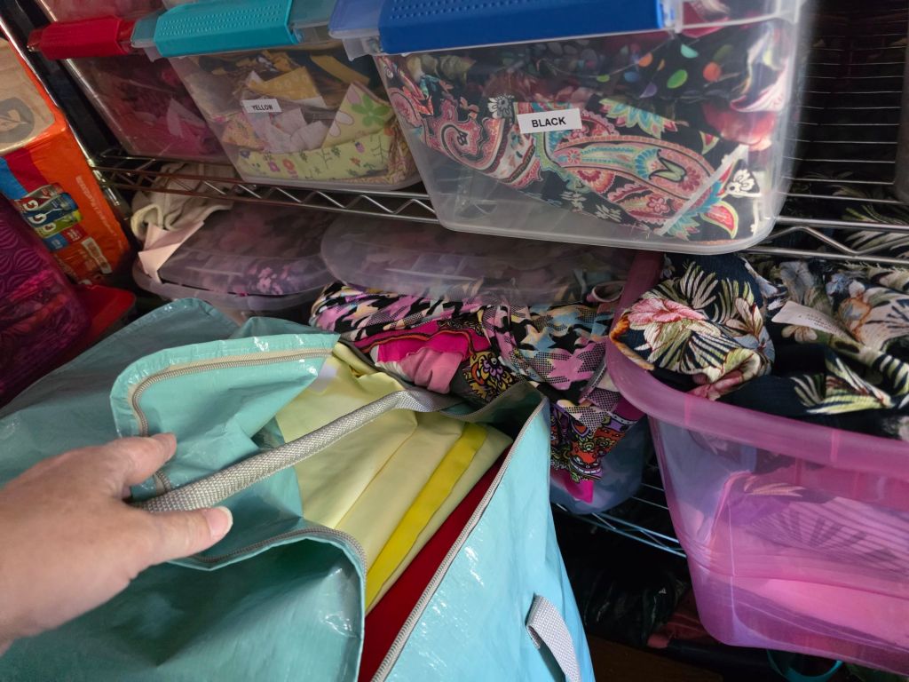 A hand reaching into a large teal bag filled with folded fabric, surrounded by colorful, labeled storage bins on a metal shelf.