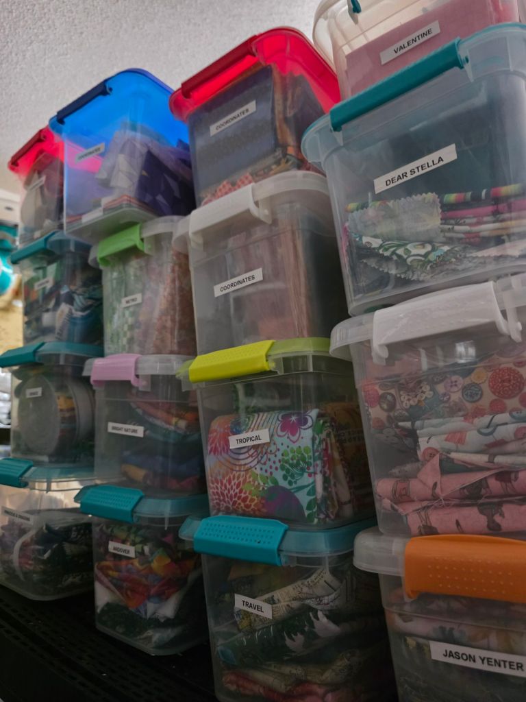 A stack of labeled storage bins containing various fabric swatches, organized by categories such as 'Coordinates', 'Tropical', 'Valentine', and 'Travel', on a shelving unit.