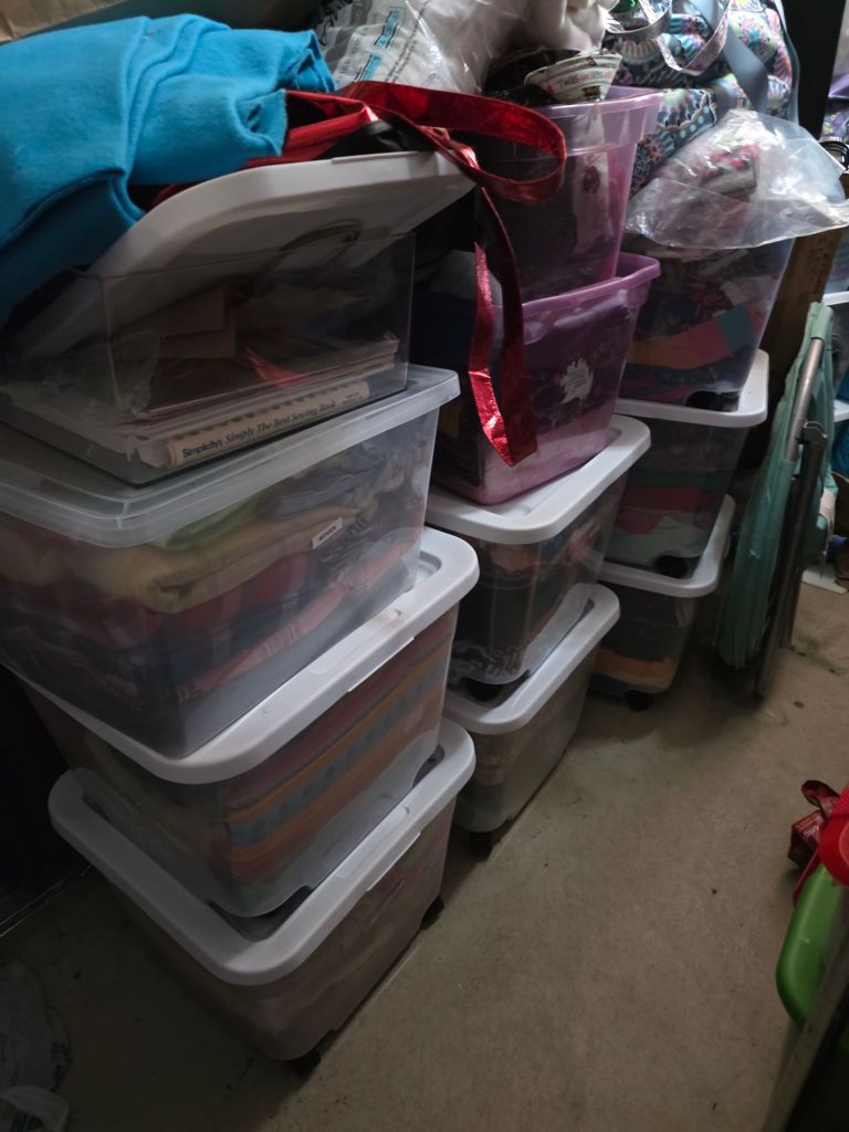 A stack of transparent storage bins filled with various items, including colorful fabrics and sewing materials, in a cluttered space.