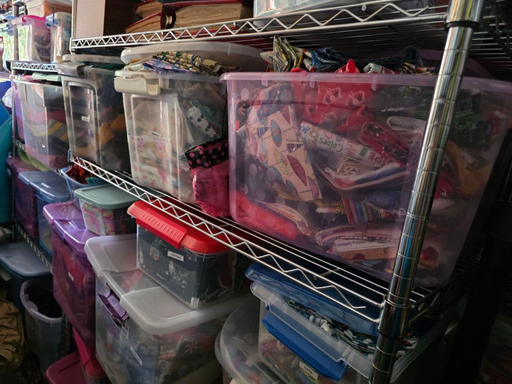 A cluttered shelving unit filled with various plastic storage bins, some clear and others colorful, containing a variety of craft materials and supplies.