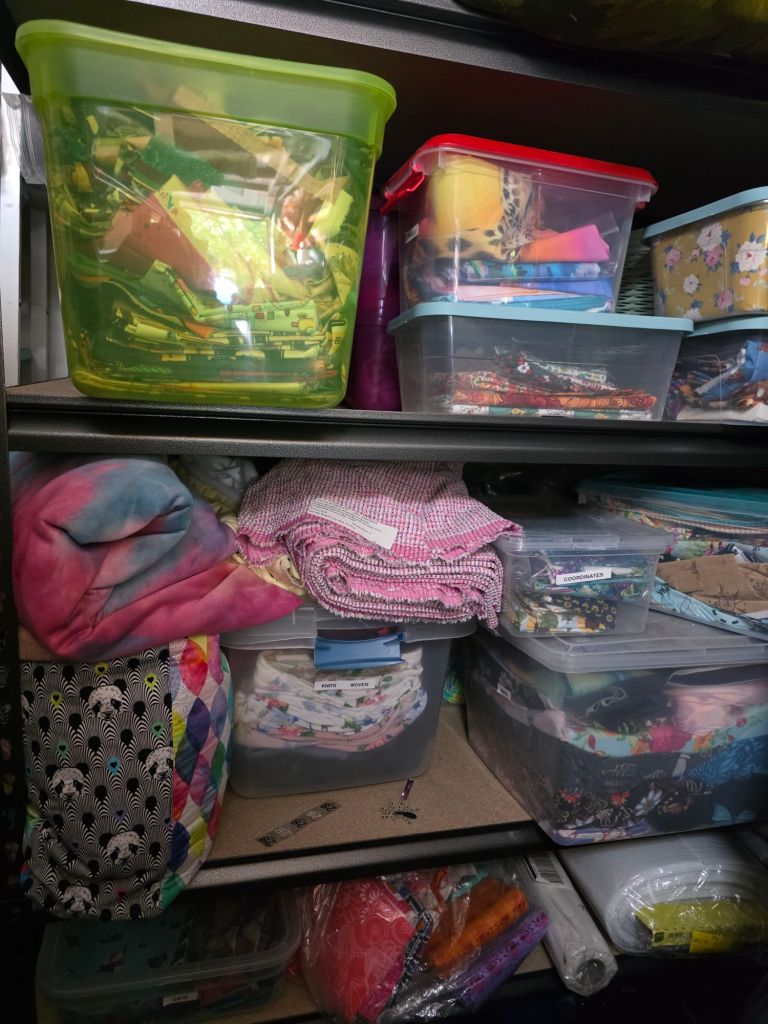 A cluttered shelf displaying various storage bins filled with craft supplies, fabric, and blankets in a range of colors and patterns.