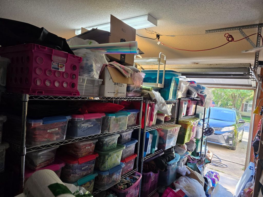 A cluttered garage interior filled with various storage bins and baskets, stacked on metal shelves, with a car visible outside in the doorway.