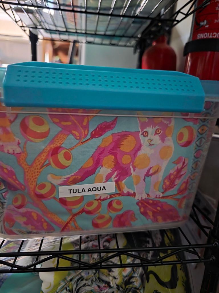 A colorful storage container featuring a vibrant design of a cat among stylized foliage and circular patterns, labeled 'TULA AQUA'.