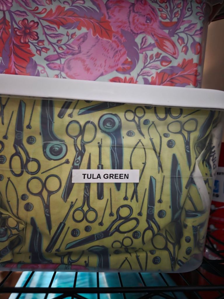A clear storage container containing fabric patterned with various sewing tools, including scissors and buttons, labeled 'TULA GREEN'.